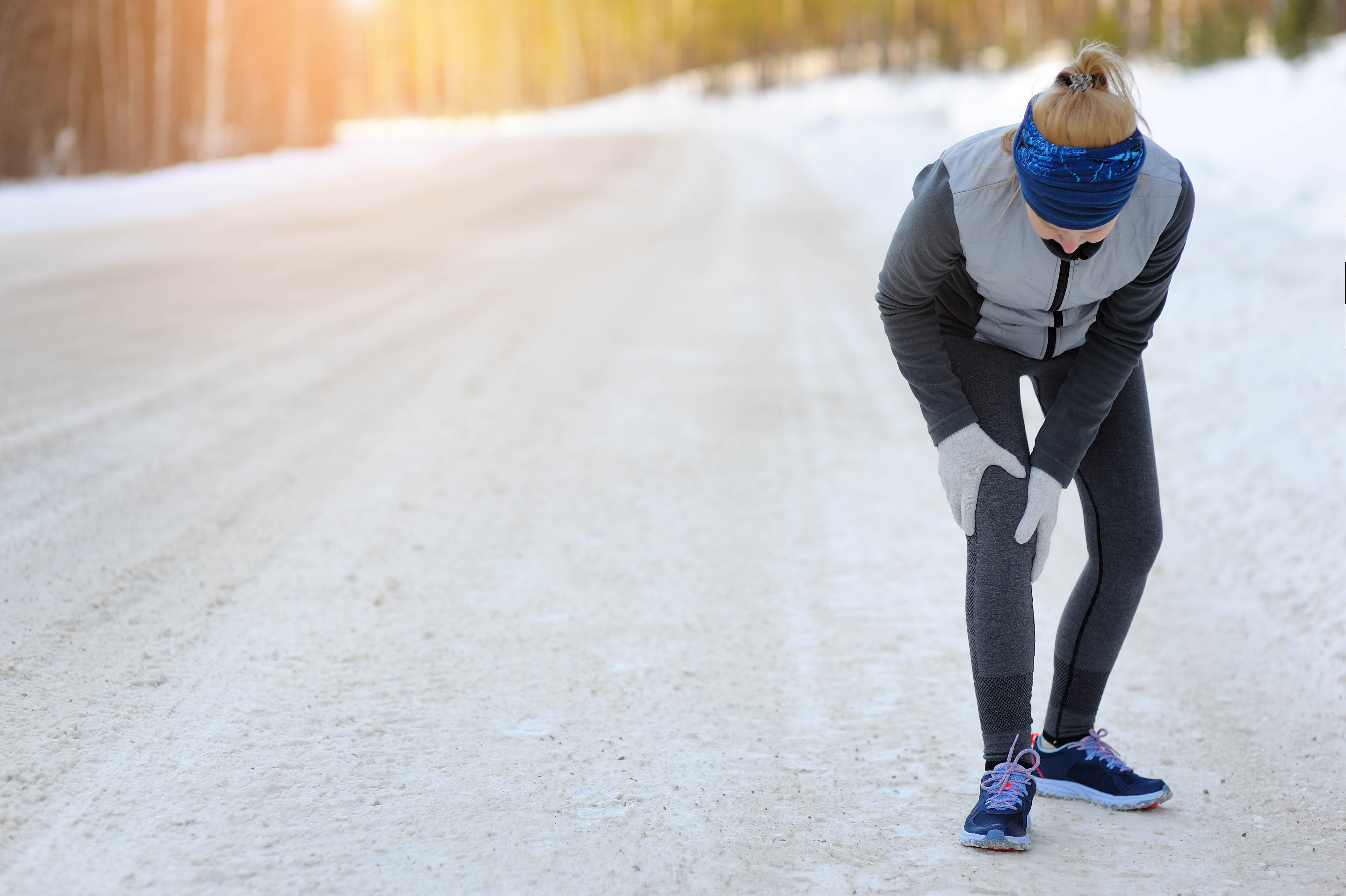 Woman wearing her winter clothing, bent over at the waist and holding her knee. Snow on the ground behind her Woman wearing her winter clothing, bent over at the waist and holding her knee. Snow on the ground behind her